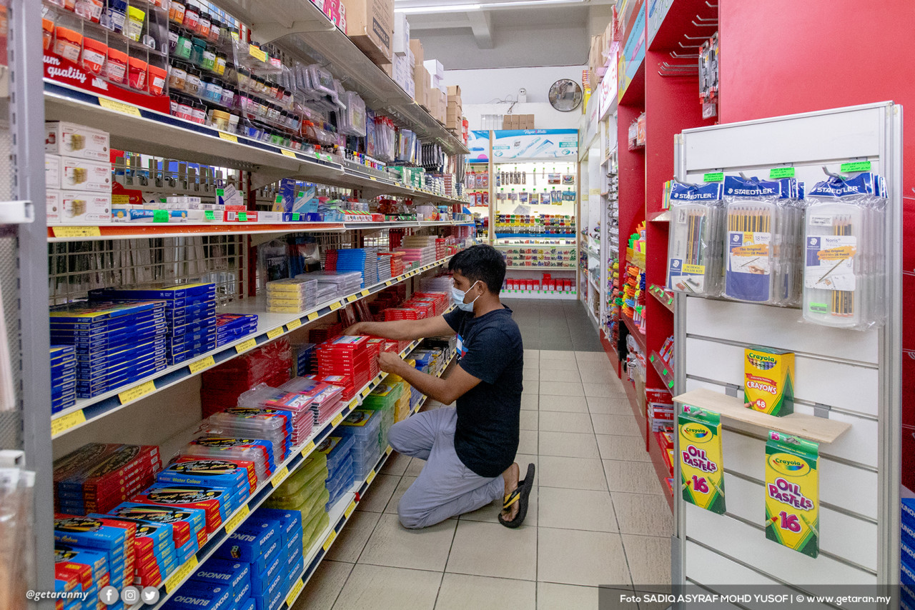 Seorang pekerja sedang menyusun barang-barang di dalam kedai alat tulis.